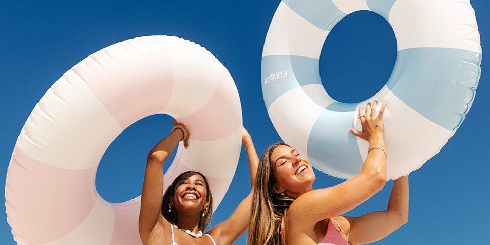 Two women in bikinis stand under a clear blue sky, smiling and holding colorful inflatable swim rings above their heads, enjoying a sunny day.