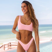 A woman in a Baby Pink Luxe Set from Honey Swim stands by a pool, smiling with long hair and jewelry, against an ocean backdrop. The clear sky enhances the tropical feel.