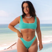A person with long dark hair stands outdoors in Honey Swims Teal Luxe Set, exuding an ocean-inspired luxury look. They wear a seashell necklace and gaze to the side with a serene ocean view as their backdrop.