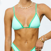 A person models the Ollie Triangle Set from Honey Swim, a mint green bikini with contrast piping, against a plain backdrop. They showcase a pearl necklace and wrist bracelets; their hair isnt visible in the image.