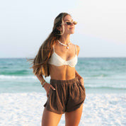 A person with long hair stands on a bright white sandy beach, smiling. They wear sunglasses, a Honey Swim white bikini top, Coconut Terry Shorts, and a necklace. The ocean and sky are in the background.