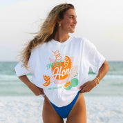On the beach, a person wears the oversized Sweet Honey Tee by Honey Swim, paired with blue bikini bottoms, earrings, and a necklace. They gaze at the vast ocean—a perfect beach cover-up moment.