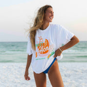 A person with long hair smiles on a beach in an oversized fit, wearing the Sweet Honey Tee by Honey Swim. The white graphic t-shirt features colorful text, paired with blue bottoms, against a stunning ocean and sky backdrop.