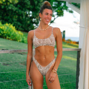 A woman smiles outdoors in the Honey Swim Rio Ribbed Bralette, her hair in a bun with jewelry. Green foliage and a white canopy are in the background.badge text: {$54 Per Piece}
