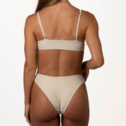 A person faces away, wearing Honey Swims Champagne Shimmer Bralette Set. The adjustable-strapped beige bikini glistens with a champagne shimmer under the lighting, as they hold the side of the bottom against a white backdrop.