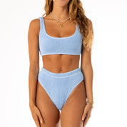 A model with wavy brown hair showcases the Honey Swim Sky Blue Luxe Set against a white backdrop, pairing its tank-style top and high-waisted bottoms with a delicate necklace and bracelets for a refined luxury look.