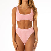 A person wearing Honey Swims Baby Pink Luxe Set poses against a white background. The scoop-neck top pairs with high-waisted bottoms. They have long wavy hair and accessorize with necklaces and bracelets.