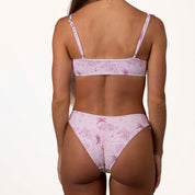 Someone is facing away, wearing a Honey Swim Positano Bralette Top in light pink with adjustable straps and a unique sketch-like pattern of palm trees and buildings against a plain white backdrop.