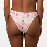 A person stands facing away in white bikini bottoms with a red Positano print of palm trees and abstract shapes. The Isla Ribbed Triangle Set from Honey Swim complements the look against a plain white background.
