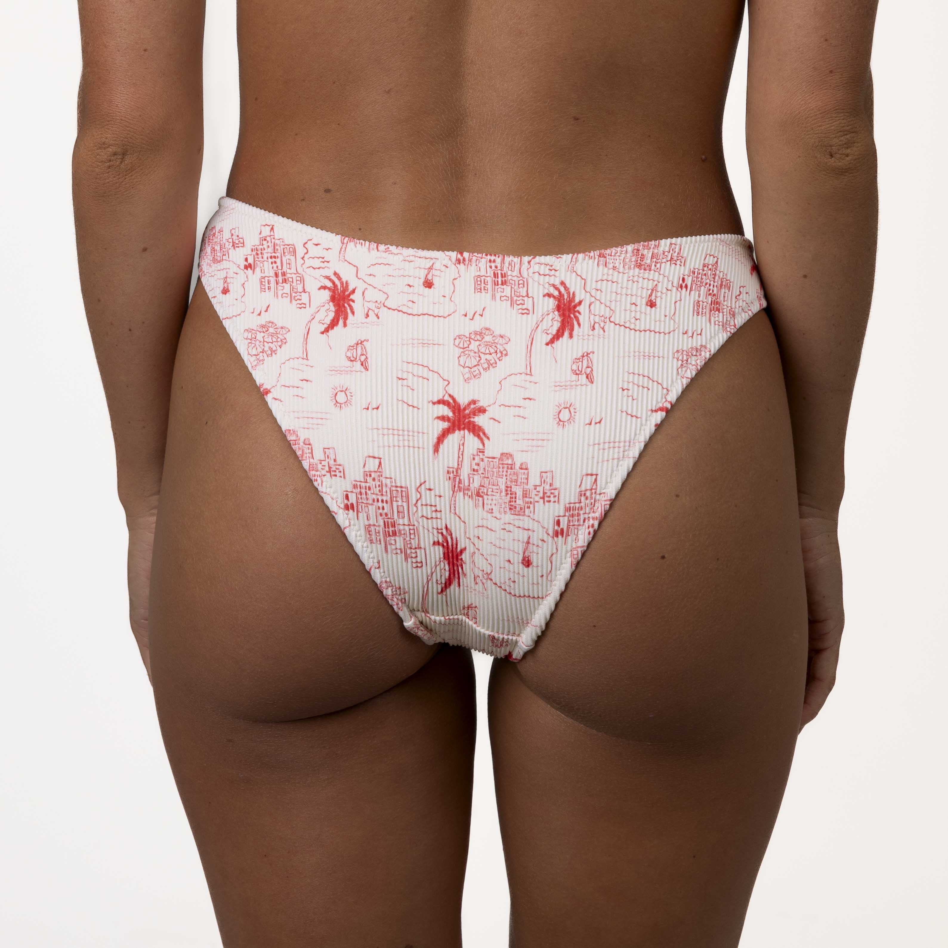 A person stands facing away in white bikini bottoms with a red Positano print of palm trees and abstract shapes. The Isla Ribbed Triangle Set from Honey Swim complements the look against a plain white background.