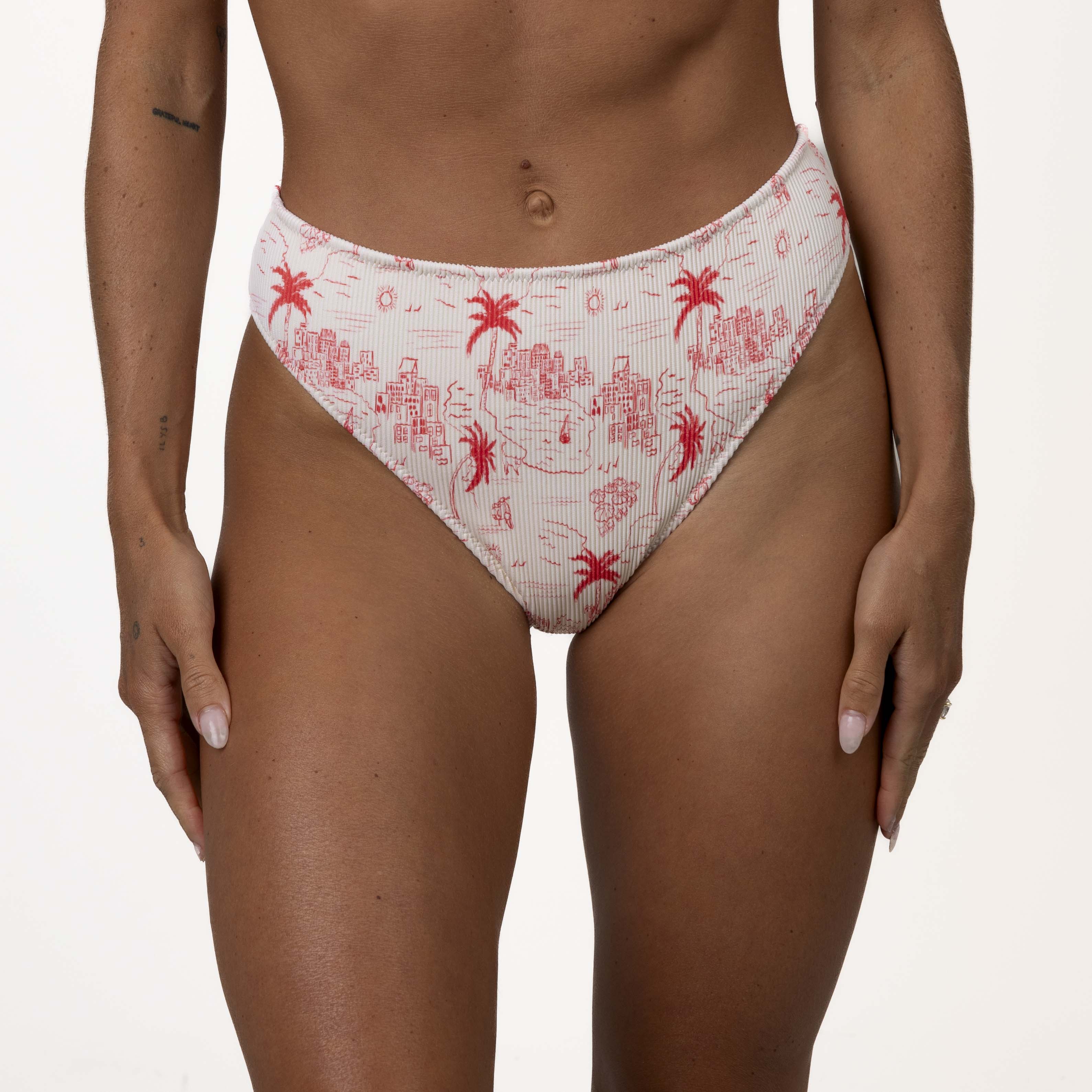A person wears Honey Swims Isla Ribbed high-waisted bikini bottoms with a red tropical print of palm trees and abstract shapes, offering supportive fit against a plain white background.