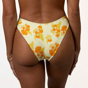 A person models the Sunni Triangle Set by Honey Swim, featuring a yellow bikini bottom with an orange floral pattern. With minimal tan lines and thin bracelets on their wrist, they pose against a plain white background.