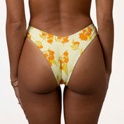 Someone models the Sunni Triangle Set from Honey Swim, showcasing the yellow bikini bottoms with an orange fruit design. Their back faces the camera, revealing gold bracelets on their left wrist against their brown skin for minimal tan lines.