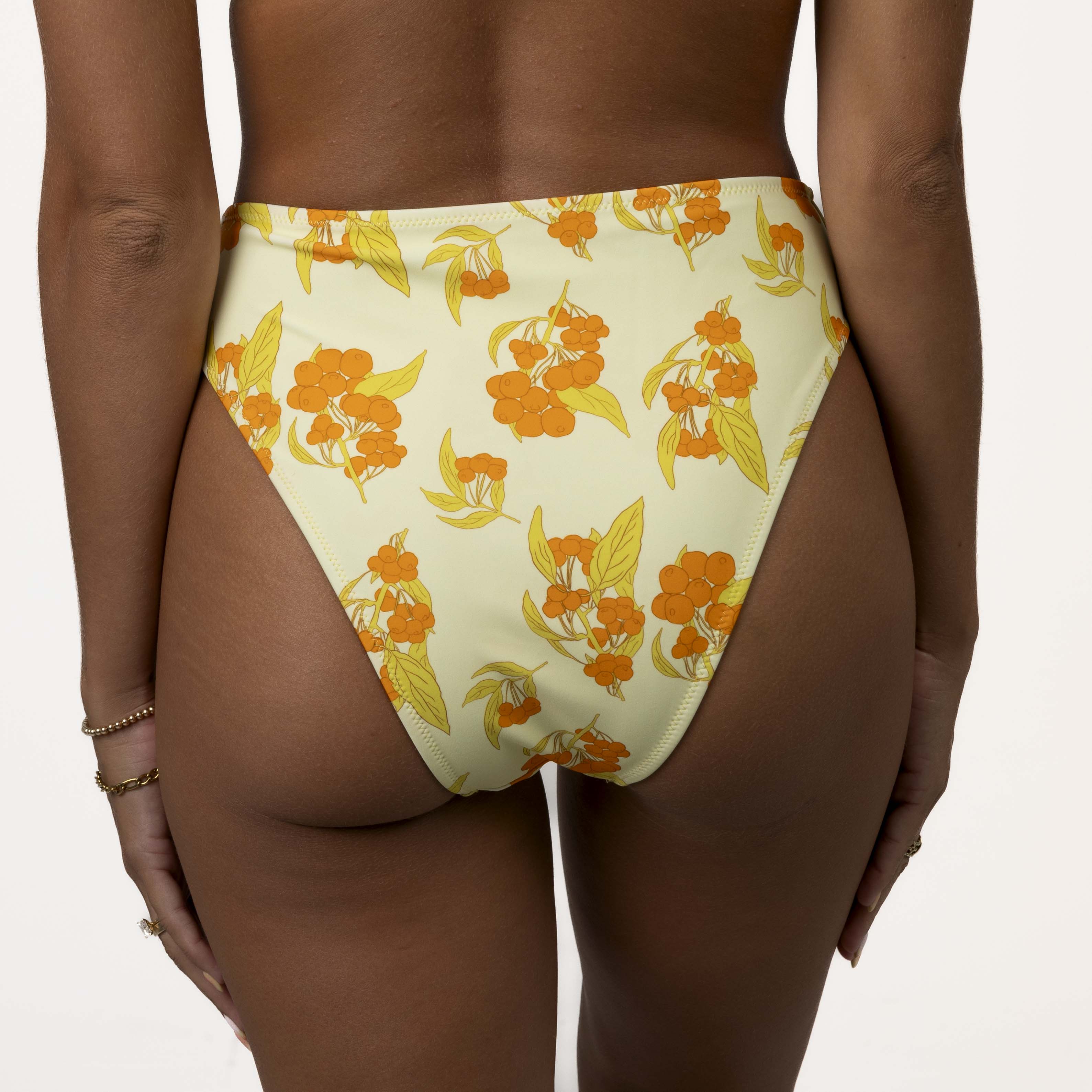 Person flaunts the Sunni High Waisted Bottoms from Honey Swim, featuring a supportive fit and moderate coverage in a yellow with orange floral pattern, paired with bracelets on one wrist against an off-white background.