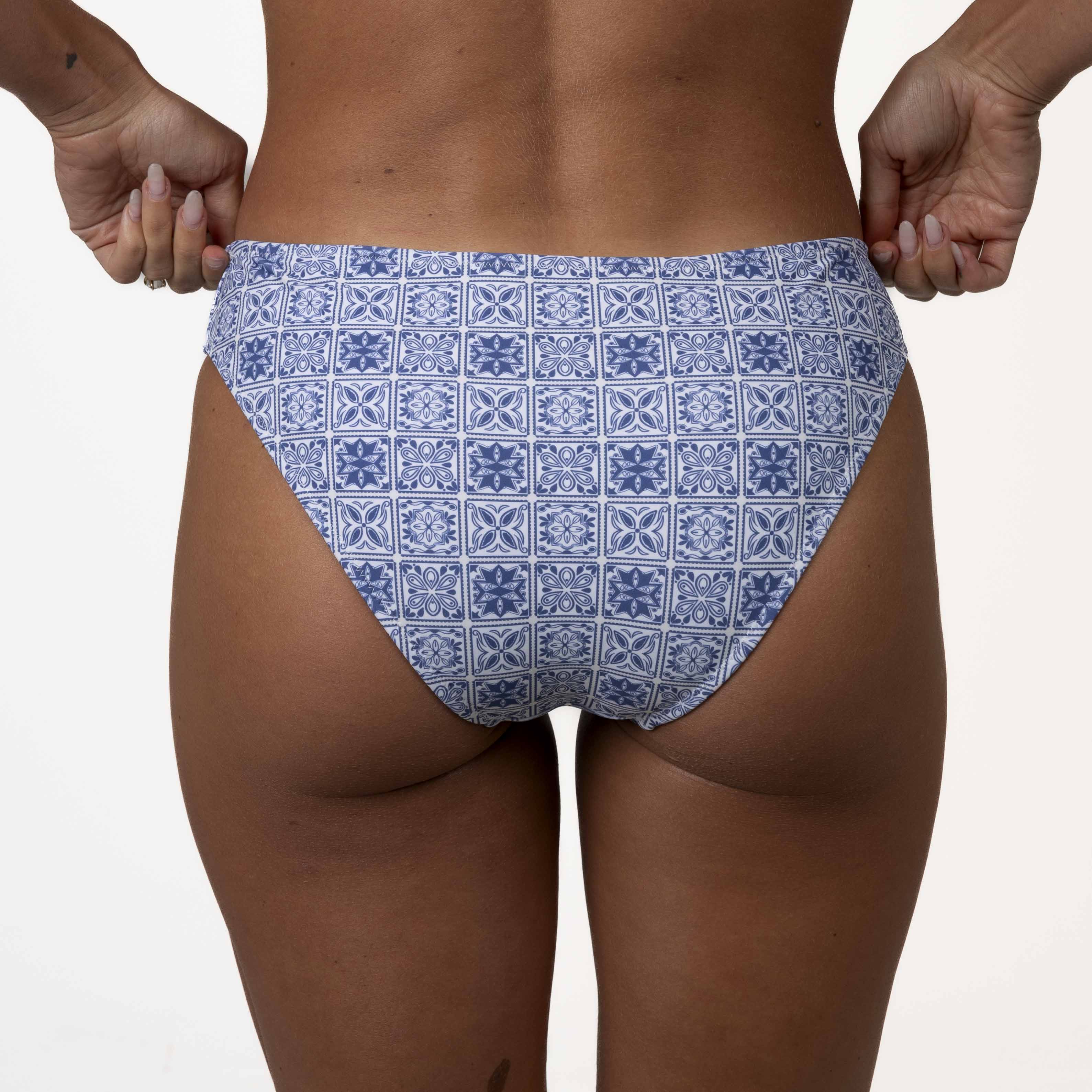 A person in a Honey Swim Mykonos Squareneck Set with a blue mosaic tile print stands against a white background, adjusting the waistband to highlight its supportive fit.