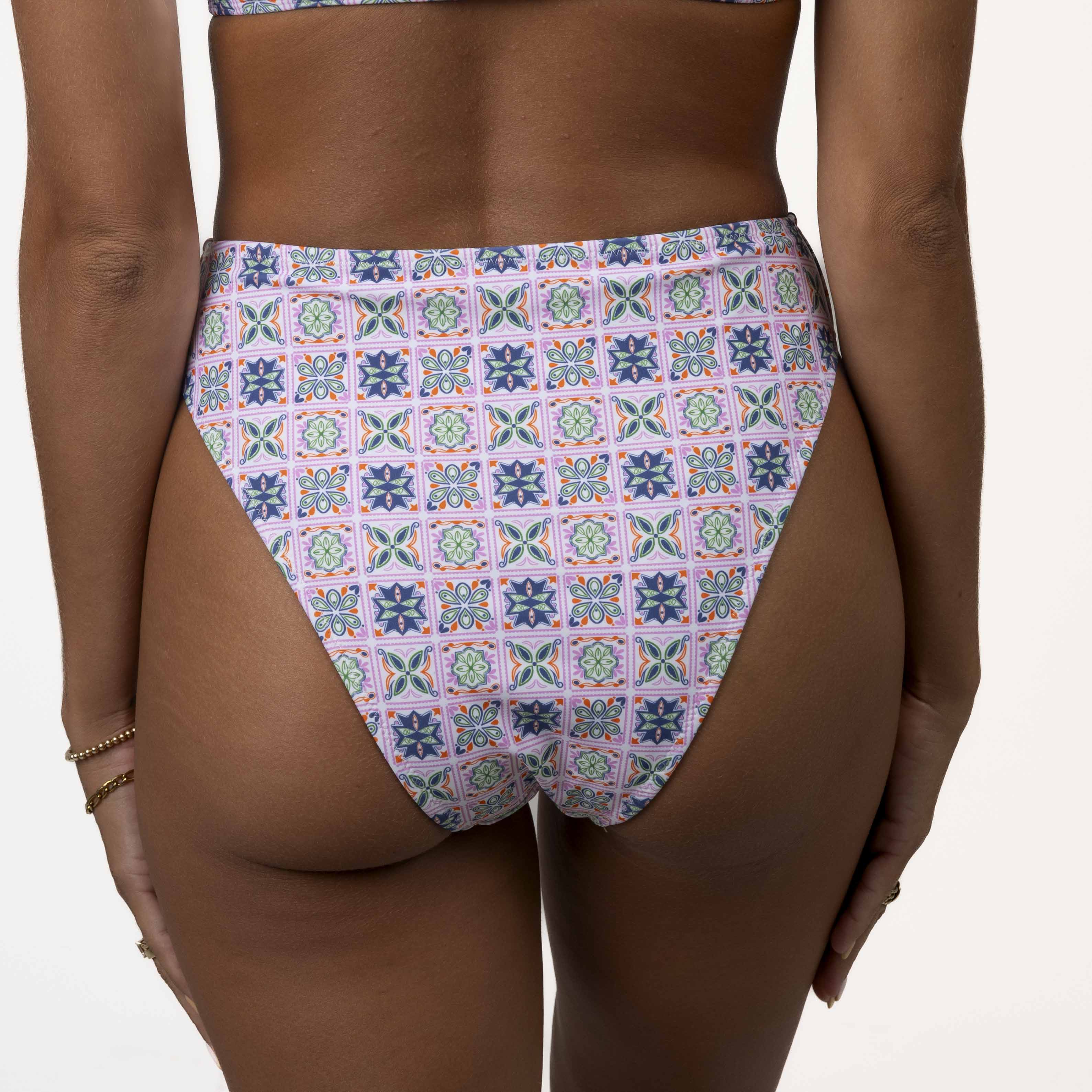 The Honey Swim Capri Bandeau Set showcases a colorful geometric pattern with floral motifs in purple, pink, and blue squares. The image highlights the lower back and bottom.
