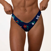 A person in Paradiso | Classic Bottoms by Honey Swim tugs the blue fabric with red, white, and green florals against a plain light backdrop, adjusting the scoop shape at the sides.