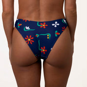 The image depicts a person from behind wearing Honey Swims Paradiso | Classic Bottoms, featuring a dark blue base with colorful floral patterns, offering moderate coverage and a flattering scoop shape against a light-colored background.