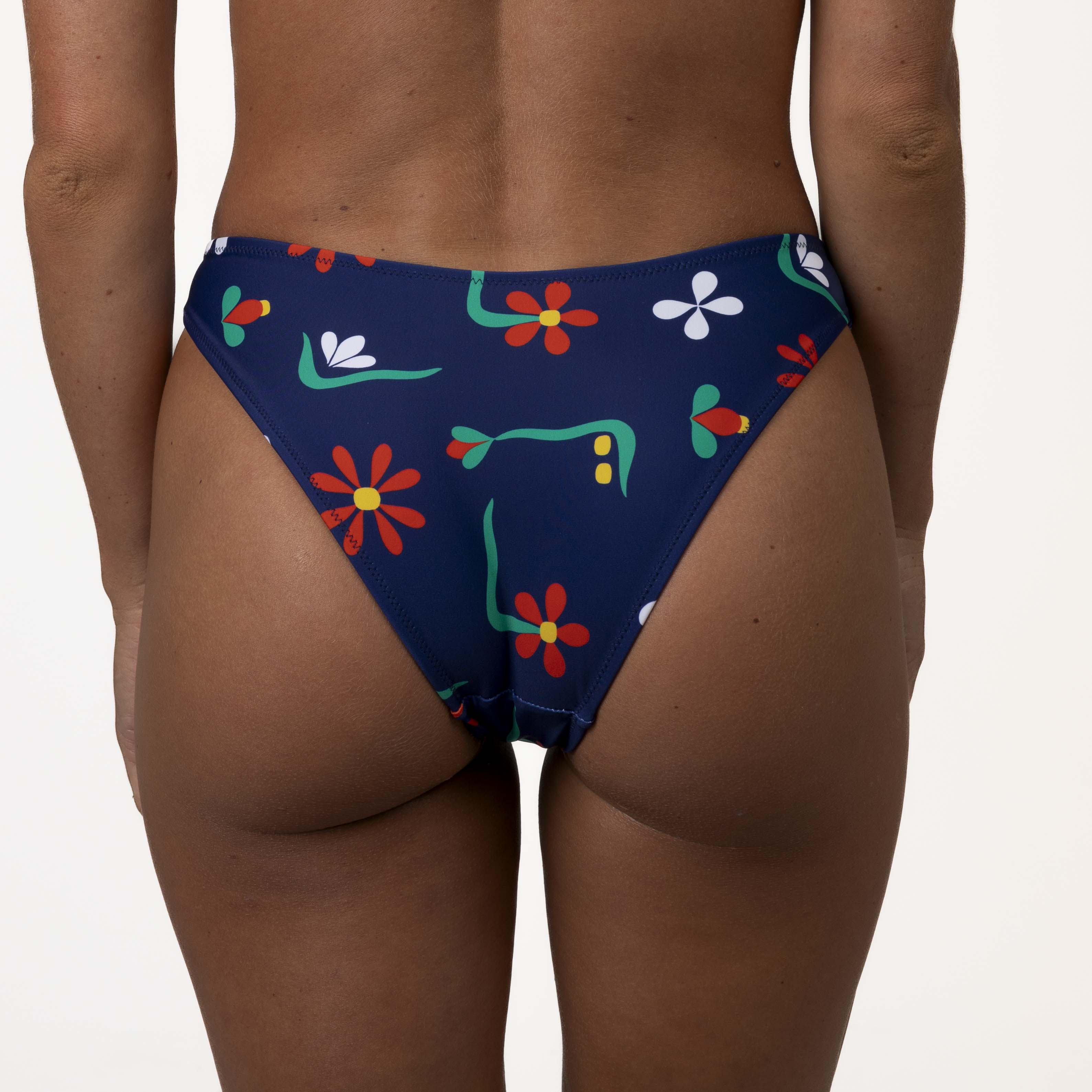 The image depicts a person from behind wearing Honey Swims Paradiso | Classic Bottoms, featuring a dark blue base with colorful floral patterns, offering moderate coverage and a flattering scoop shape against a light-colored background.