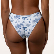 Person wearing Honey Swims Amalfi | Classic Bottom, featuring a white bikini bottom with blue toile patterns of trees, houses, and bicycles. The scoop shape offers moderate coverage, embodying the timeless Amalfi style.