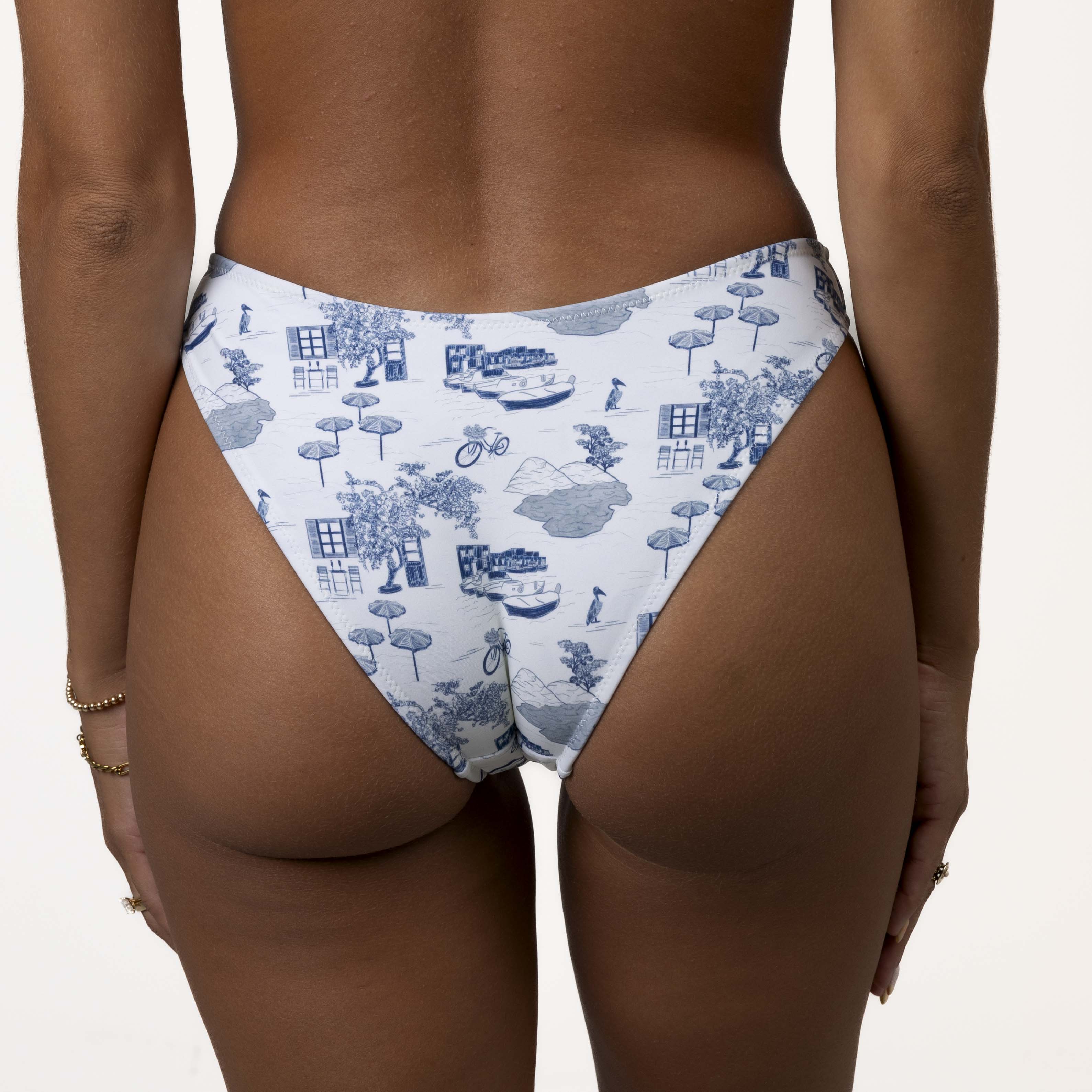 Person wearing Honey Swims Amalfi | Classic Bottom, featuring a white bikini bottom with blue toile patterns of trees, houses, and bicycles. The scoop shape offers moderate coverage, embodying the timeless Amalfi style.