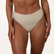 A person in Honey Swims Champagne Shimmer Bandeau Set and beige, high-waisted bikini bottoms is shown from waist to mid-thigh, with a tattoo on their left forearm, against a plain white background.