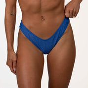 A person wearing Honey Swims Azure Shimmer Bralette Set holds the sides of their blue bikini bottom against a plain white wall.