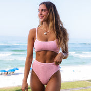 On a grassy area by the beach, a person wears the Honey Swim Baby Pink Sporty Luxe Set. Theyre smiling with flowing long hair and a beaded necklace as the sea and umbrellas appear in the background.