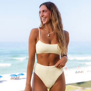 A woman with long hair smiles on a beach, wearing the Honey Swims Honey Sporty Luxe Set and a pearl necklace. The scene includes a clear sky, ocean waves, and distant beach umbrellas.