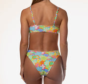 A rear view showcases a person in a vivid Honey Swim Eden Bralette bikini against a plain white background. The tropical pattern pairs beautifully with their shoulder-length hair and bracelets, creating a timeless silhouette.