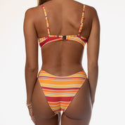 Wearing the Honey Swim Sunset Underwire Set, a person stands against a plain light background. The vibrant orange, red, and yellow stripes capture light beautifully, showcasing the bikini’s seamless comfort from the rear view.