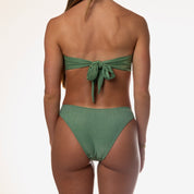 The woman is wearing an Olive Shimmer Bandeau Set by Honey Swim. The strapless tie-back bikini top forms a bow at her back, and she wears a bracelet on her left arm against a plain white background.