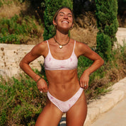 A smiling woman in a light pink Positano Bralette by Honey Swim, with adjustable straps, stands outdoors. Shes wearing gold jewelry, hands on hips, amid greenery and a stone path.