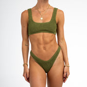 Wearing the Olive Luxe Set by Honey Swim, a person showcases an olive green knitted bikini featuring a square-neck top and high-waisted bottoms, enhanced by gold necklaces and bracelets against a pristine white background.