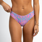 A person wearing Honey Swims Starfish, full coverage swim bottoms in a whimsical pink and blue pattern, adjusts them against a plain white background. They wear a gold bracelet and ring for an elegant touch to the playful look.
