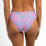A person wears Honey Swims Starfish full coverage bottoms in a whimsical multicolored abstract pattern, paired with thin bracelets on their left wrist, against a white background with their back to the camera.