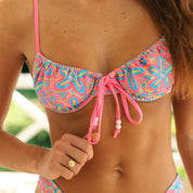 A person in the Starfish Underwire Top by Honey Swim, featuring a colorful pink and blue pattern, adjusts the front tie. They wear a gold ring, with blurred greenery in the background.