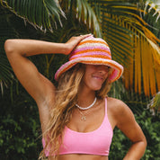 Someone in a pink top poses against lush greenery, wearing Honey Swims vibrant Hibiscus Hat and smiling. Accessorized with a pearl necklace, their long hair complements the perfect beach day vibe.