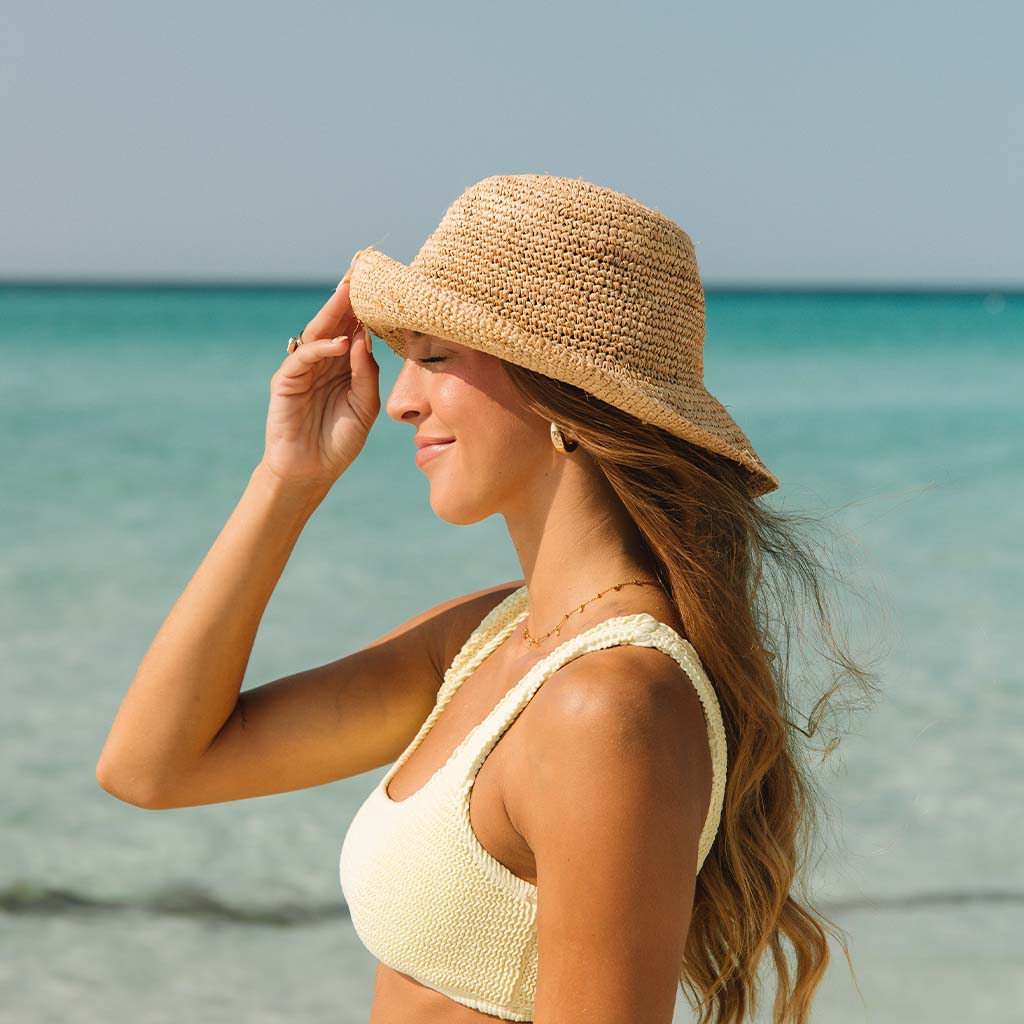 A woman in a Honey Swim beige bikini top and The Inlet hat stands on a sunlit beach, gazing at the sea. She holds her adjustable hats brim as the ocean sparkles under the bright sky.
