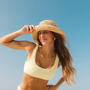A woman in a yellow bikini and beach hat smiles under the blue sky. She has long brown hair, holding her adjustable Honey Swim The Inlet hat, ready for travel adventures.