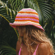 A person with long wavy hair is in profile, wearing a wide-brimmed striped Hibiscus Hat by Honey Swim in pink, orange, and cream. Theyre dressed in a pink top and beaded necklace against lush tropical leaves—perfect for a beach day.