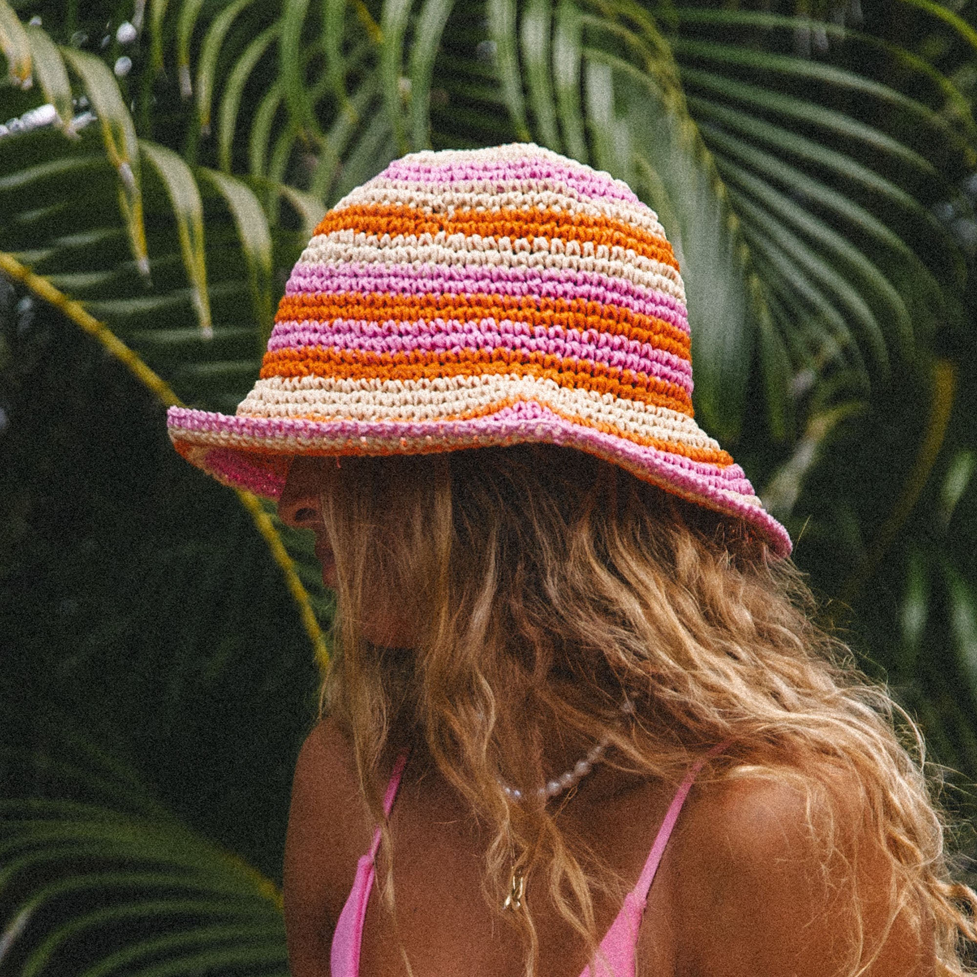 A person with long wavy hair is in profile, wearing a wide-brimmed striped Hibiscus Hat by Honey Swim in pink, orange, and cream. Theyre dressed in a pink top and beaded necklace against lush tropical leaves—perfect for a beach day.
