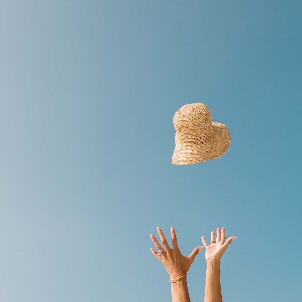 Hands reaching upwards as The Inlet Hat by Honey Swim, featuring an adjustable interior strap, is tossed into a clear blue sky.