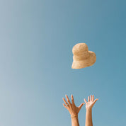 Hands reaching upwards as The Inlet Hat by Honey Swim, featuring an adjustable interior strap, is tossed into a clear blue sky.