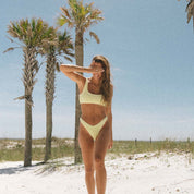 On a sandy beach, a woman in a Honey Luxe Set by Honey Swim shields her eyes from the sun. Three palm trees frame the background beneath a clear blue sky, enhancing her luxurious style.