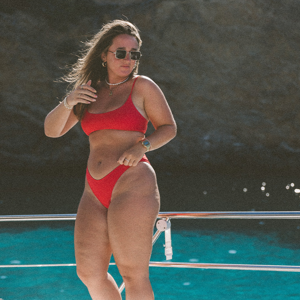 On a boat under the sun, a woman in honeyswims Recycled Flat Red bikini bottom stands against turquoise water and rocky cliffs. She wears sunglasses and jewelry as she adjusts her hair.