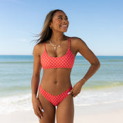 A woman in the sporty, flattering Honey Swim Azalea Bralette Set with adjustable straps stands on a sandy beach, smiling with the ocean and blue sky in the background.