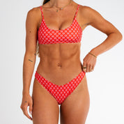 A woman models the Honey Swim Azalea Bralette Set, a sporty red bikini with white floral print and adjustable straps, posing against a white background with her right hand on her bikini bottom.