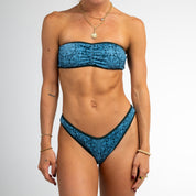 A woman models the Honey Swim Bay Ribbed Cinched Bandeau Top in blue, styled with layered necklaces and bracelets, standing against a white background. Face not visible. Perfect for swimwear inspiration.