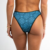 Model wearing Honey Swim’s Bay Ribbed bikini bottoms, featuring a blue pattern, shown from the back with bracelets on both wrists against a plain white background.